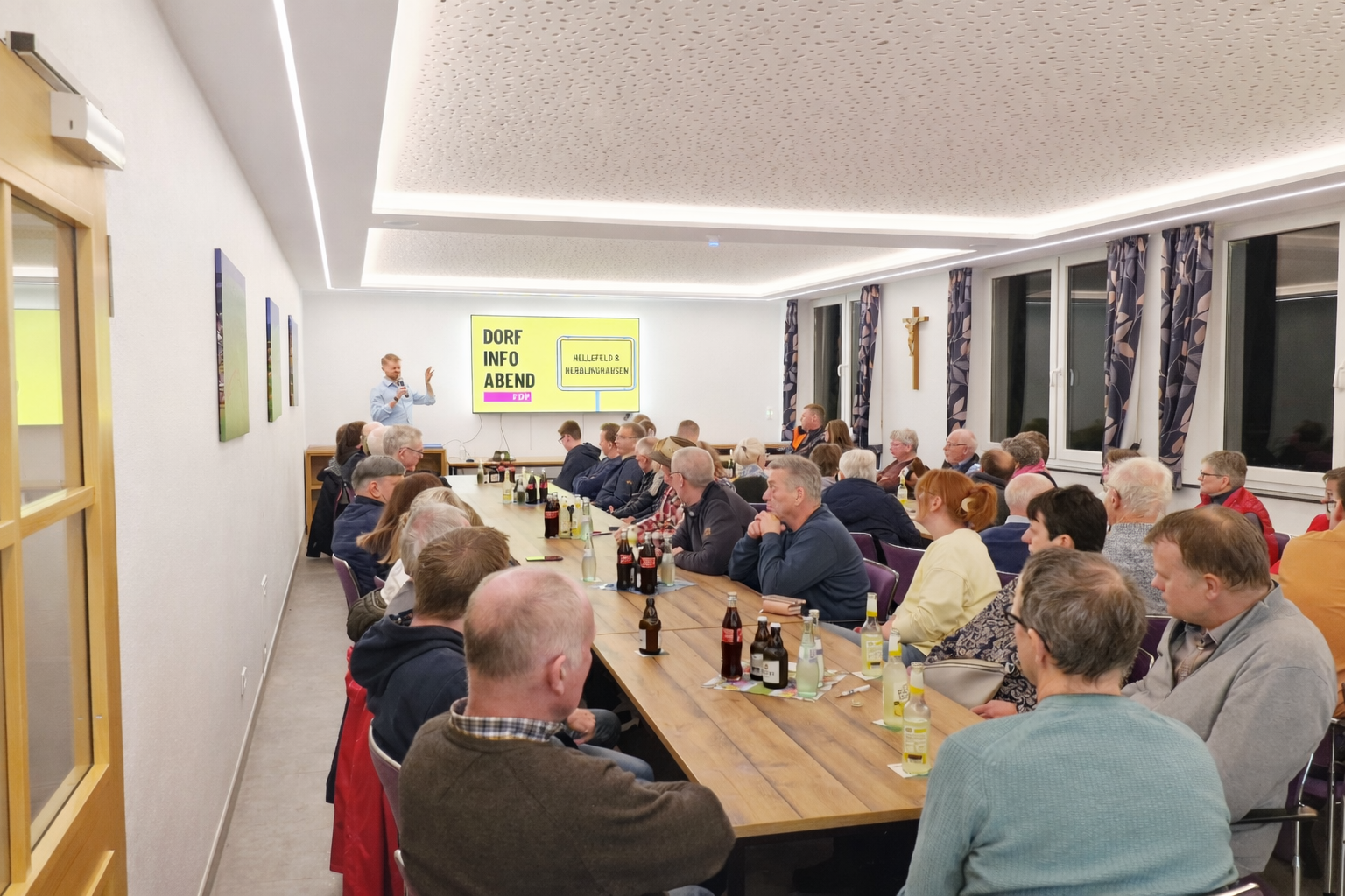 Gut besuchter Dorf-Info-Abend in modernem Veranstaltungsraum: Tom Rengshausen präsentiert vor rund 30 Zuhörern an einer Leinwand, die Gäste sitzen aufmerksam an einer langen Tafel.