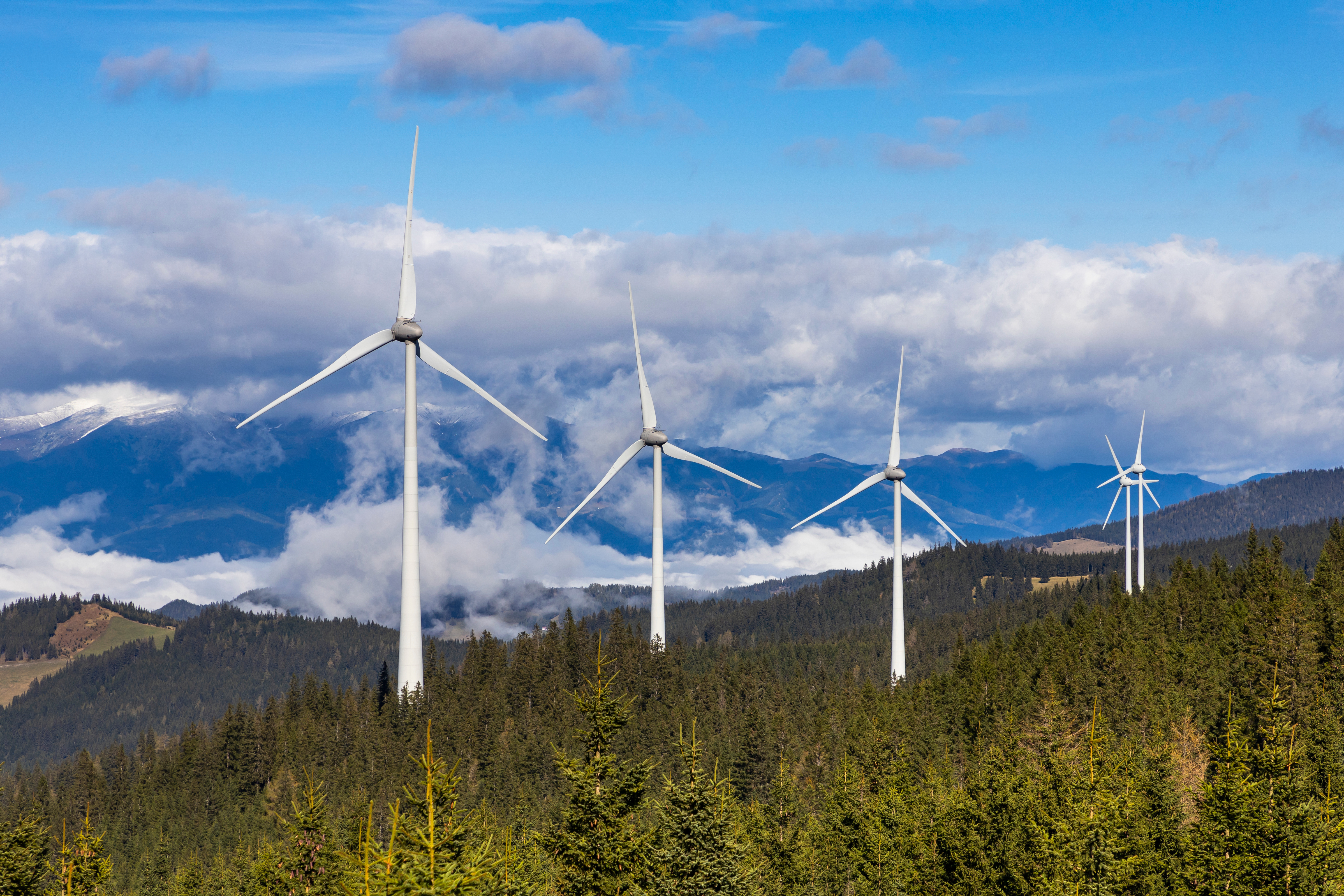 Windkraftanlagen auf bewaldeten Hügeln in einer Mittelgebirgslandschaft, mit Wolken und Bergen im Hintergrund.