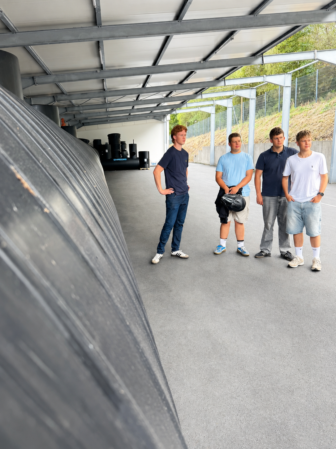 Vier junge Männer der Jungen Liberalen stehen während einer Betriebsführung in einer überdachten Produktions- und Lagerhalle von ENREGIS. Sie blicken aufmerksam nach rechts in Richtung des nicht sichtbaren Gesprächspartners. Die gesamte linke Bildhälfte wird von einer sehr großen schwarzen, gerippten Anlage eingenommen, die vom unmittelbaren Vordergrund weit in die Tiefe der Halle reicht. Weitere schwarze Anlagen und Bauteile sind im Hintergrund zu sehen. Der Größenvergleich mit den vier Personen verdeutlic