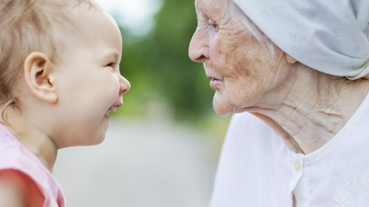 Ein Kleinkind strahlt vor einem ländlichen Hintergrund, vielleicht ein Bauerhof, einen Menschn mit einem recht alten und faltigen Gesicht an.