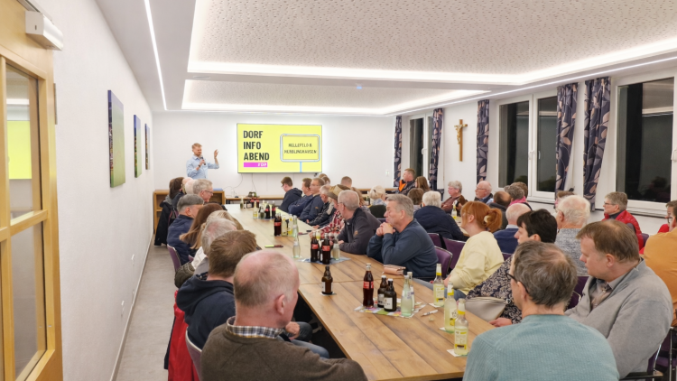 Gut besuchter Dorf-Info-Abend in modernem Veranstaltungsraum: Tom Rengshausen präsentiert vor rund 30 Zuhörern an einer Leinwand, die Gäste sitzen aufmerksam an einer langen Tafel.