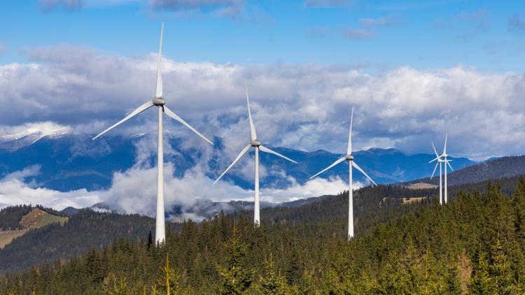 Windkraftanlagen auf bewaldeten Hügeln in einer Mittelgebirgslandschaft, mit Wolken und Bergen im Hintergrund.