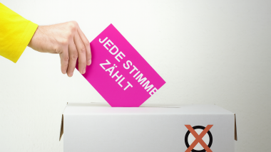 Eine Hand steckt eine Wahlkarte in eine Wahlurne. Auf der Karte steht in Großbuchstaben: „JEDE STIMME ZÄHLT“.