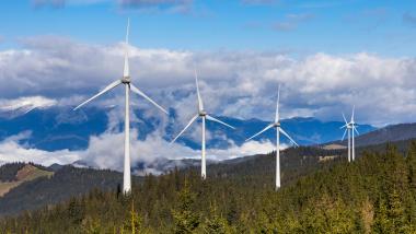 Windkraftanlagen auf bewaldeten Hügeln in einer Mittelgebirgslandschaft, mit Wolken und Bergen im Hintergrund.